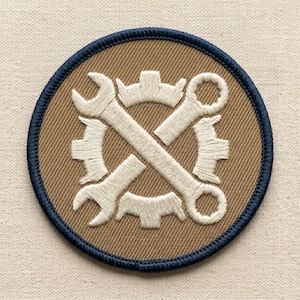 May include: Embroidered patch featuring a crossed wrench and gear design in white on a tan background. The patch has a navy blue border and is circular in shape. The design suggests a mechanical or engineering theme.