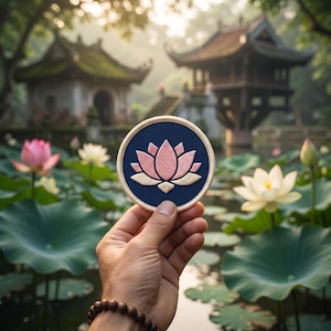 May include: A circular embroidered patch featuring a stylized pink and cream lotus flower design on a dark blue background, finished with a light cream stitched border. The patch is held up against a backdrop of a pond with blooming lotus flowers.