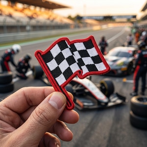 May include: A small embroidered patch shaped like a checkered flag with a bold red border. The flag features a classic black and white square pattern, with the fabric edges stitched in red thread to create a distinct outline for the design.