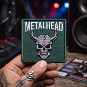 May include: A square embroidered patch with a dark green background featuring the white text METALHEAD at the top and a central silver-toned skull graphic with two curved horns. The patch has a dark green stitched border and a flat square shape.