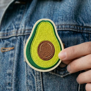 May include: Embroidered avocado patch featuring a green and yellow design with a brown seed. The patch is attached to a denim jacket. The avocado patch is a fun accessory.