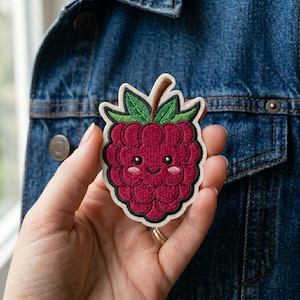 May include: Embroidered patch featuring a smiling raspberry with green leaves and a brown stem. The raspberry is a deep red color with a white border. The patch is held in front of a blue denim jacket.