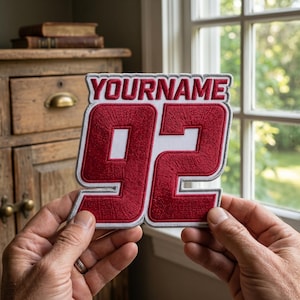 May include: A rectangular patch with the text "YOURNAME" above the number "92". The numbers and text are in a bold, red, embroidered font with a white border and a gray outline. The patch is held in front of a window.
