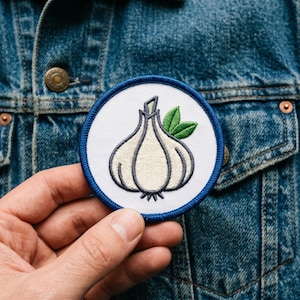 May include: A round embroidered patch featuring a garlic bulb design with green leaves. The patch has a white background with a blue border. The patch is being held in front of a denim jacket.