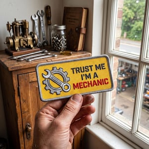 May include: A yellow patch with the text "TRUST ME I'M A MECHANIC" and a wrench and gear graphic. The patch is held in front of a wooden cabinet with tools and a small engine model.