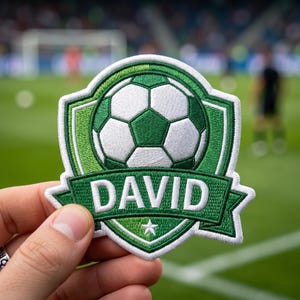 May include: Embroidered patch featuring a soccer ball design in shades of green and white. The name "DAVID" is displayed in white lettering on a green banner. A small white star is at the bottom. The patch has a white border.