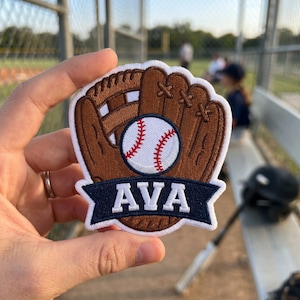 May include: A baseball-themed patch featuring a brown baseball glove with a white baseball and red stitching. A navy blue banner at the bottom displays the name "AVA" in white letters. The patch is likely for a baseball team or player.