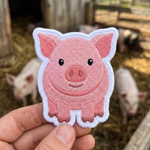 May include: An embroidered patch featuring a cartoon pig with light pink skin, dark pink ears, and a snout. The pig has small black eyes and a simple line for a mouth. The patch has a thick white border that follows the outline of the pig's shape.