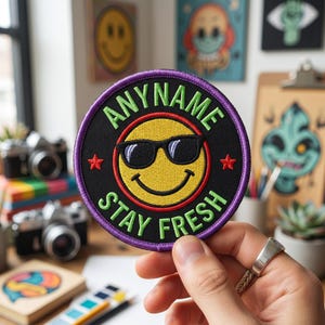 May include: A circular embroidered patch with a black background and a purple border. The center features a yellow smiley face wearing black sunglasses. The text ANY NAME is arched above and STAY FRESH is arched below in light green letters.