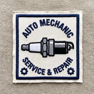 May include: Square patch with a white background and a navy blue border. The patch features the words "AUTO MECHANIC SERVICE & REPAIR" in navy blue, surrounding a detailed illustration of a spark plug. Two gear icons flank the text.