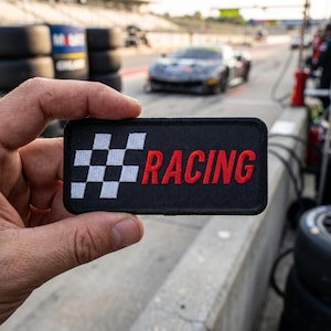 May include: A rectangular black embroidered patch featuring a white checkered flag pattern on the left side and the word RACING in bold red capital letters on the right side, with a black stitched border surrounding the entire design.