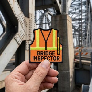May include: An orange safety vest patch with yellow reflective stripes and a black border. The patch features the text BRIDGE INSPECTOR in bold black letters and a small vertical ruler graphic on the right side, all set against a black background.