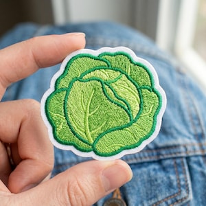 May include: Embroidered patch featuring a green cabbage design with detailed stitching. The cabbage is surrounded by a white border. This patch is suitable for clothing, bags, or other fabric items.