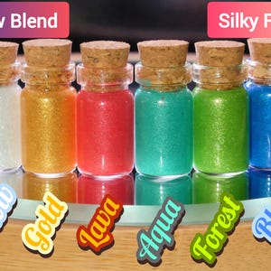 May include: Six small glass bottles with cork stoppers, each filled with a different color of glitter. Colors include white, gold, red, teal, green, and blue. Text on the image reads "New Blend" and "Silky Feel!" with color names below each bottle.