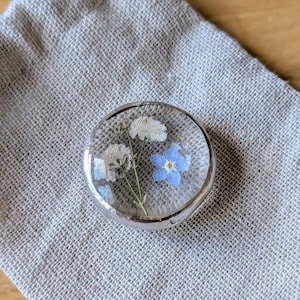 May include: A clear, circular resin paperweight featuring dried flowers, including a blue forget-me-not and white blossoms, encased in a silver-coloured metal frame. The paperweight sits on a beige linen pouch, evoking a natural, botanical aesthetic.