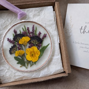 Könnte beinhalten: Ein kreisförmiges Harzornament mit gepressten gelben Blumen und violetten Zweigen in einem silberfarbenen Metallrahmen mit einem hellvioletten Samtband. Es liegt in einer Schachtel mit einer Karte, auf der steht: YOU ARE IN MY thoughts.