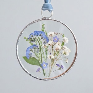 Könnte beinhalten: Ein kreisförmiges Glasornament mit silbernem Metallrahmen, das gepresste blaue und weiße Blumen mit grünem Blattwerk enthält. Das Ornament hängt an einem hellblauen Samtband und zeigt ein zartes Arrangement aus getrockneten botanischen Elementen.