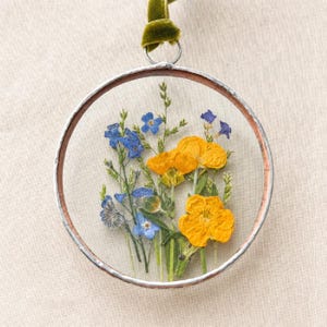 Puede incluir: Un adorno circular de vidrio enmarcado en un borde metálico fino que contiene flores de ranúnculo amarillas prensadas y pequeñas flores de nomeolvides azules con tallos verdes. Una cinta de terciopelo verde se sujeta a una anilla metálica.