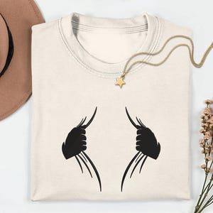 May include: Cream-colored t-shirt with a black graphic design. The design features two stylized claw-like shapes. A gold necklace with a star pendant rests on the shirt. A brown hat and dried flowers are also in the image.
