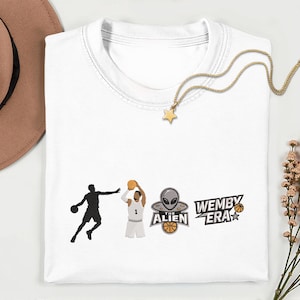 May include: White t-shirt with basketball-themed graphics. Features a silhouette of a player dribbling, another shooting, and an alien with a basketball. Text reads "WEMBY ERA". A gold star pendant necklace rests on the shirt.