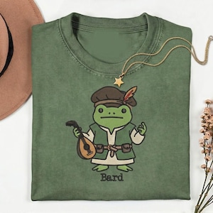 May include: A green t-shirt featuring an embroidered cartoon frog dressed as a bard, holding a lute. The frog wears a brown hat with a feather and a green vest. The word "Bard" is printed below the frog.