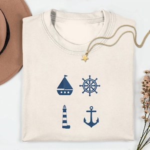 May include: Cream-colored t-shirt featuring navy blue nautical icons: a sailboat, ship's wheel, lighthouse, and anchor. A gold star pendant necklace rests on the shirt. A brown hat and dried flowers are also in the image.