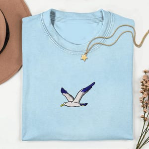 May include: A light blue t-shirt with a crew neck featuring an embroidered seagull in flight. The seagull has white and gray feathers with blue wingtips and a yellow beak. A gold star pendant necklace rests on the shirt.