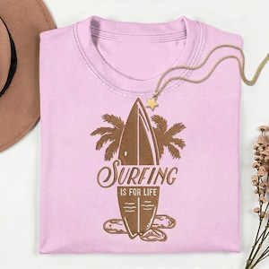 May include: A light pink t-shirt with a brown surfboard graphic and the text "Surfing is for Life." A gold necklace with a star pendant rests on the shirt. A brown hat is visible in the upper left corner.