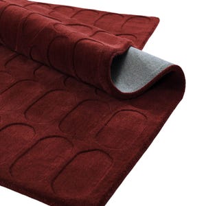May include: A deep red rectangular rug featuring a repeating embossed pattern of rounded rectangles or pill shapes across its surface. The rug is partially folded over, revealing a smooth, light gray backing material on the underside of the fabric.