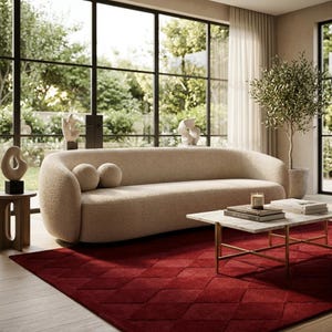 May include: A curved beige bouclé sofa with two round matching cushions, positioned in a room with a deep red diamond-patterned rug, a white marble coffee table with gold legs, and a small wooden side table holding a sculptural object.