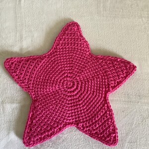May include: A handmade, bright pink crocheted star. The star is made with a spiral pattern, and the edges are well-defined. The star is a decorative item, possibly a coaster or trivet, and is placed on a white surface.