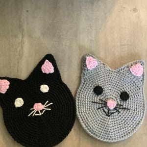 May include: Two crocheted cat-shaped coasters. One is black with pink ears, white eyes, and a pink nose with white whiskers. The other is gray with pink ears, black eyes, and a pink nose with black whiskers. Both are on a light brown surface.