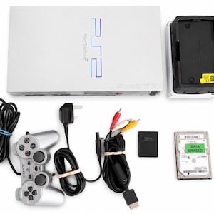 Könnte beinhalten: Eine weiße PlayStation 2 Konsole mit blauem Schriftzug, ein silberner Controller, ein schwarzes Netzkabel, ein mehrfarbiges AV-Kabel, eine schwarze Speicherkarte, eine Festplatte mit grünem DATA ERASED Aufkleber und ein schwarzer Standfuß mit gelbem Warnetikett.