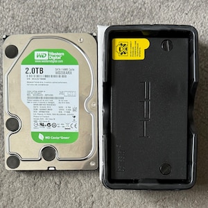 Może przedstawiać: Dysk twardy Western Digital 2,0 TB z zieloną etykietą i czarną obudową GameStar. Dysk twardy zawiera tekst "WD Caviar Green" i inne specyfikacje techniczne. Obudowa jest czarna z logo GameStar.