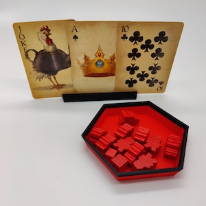 Op de afbeelding: Een unieke kaartspelset met drie oversized speelkaarten met vintage illustraties, waaronder een haan, een kroon en de tien van klaveren. Een rode bak met houten spelstukken staat onder de kaarten.