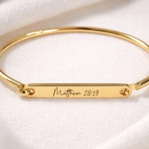 May include: A gold-colored bangle bracelet with a rectangular bar in the center. The bar is engraved with the text "Matthew 28:19". The bracelet is set against a white, silky background.