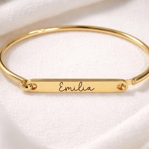 May include: A gold-colored bangle bracelet with a rectangular bar in the center. The bar is engraved with the name "Emilia" in cursive script. The bracelet is displayed on a white, silky surface.