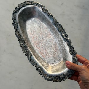Op de afbeelding: Een ovaal zilverkleurig metalen dienblad met een ingewikkeld reliëf bloemenpatroon in het midden en een decoratieve geschulpte rand met gedetailleerd krulwerk en bladmotieven langs de gehele buitenrand van de schaal.