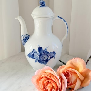 May include: A white porcelain coffee pot with a blue floral design. The pot has a lid and a curved handle. Two peach-colored roses are in the foreground. The pot is on a white marble surface.