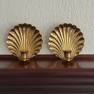 May include: A pair of matching gold-toned brass wall sconces, each featuring a large, fan-shaped scallop shell backplate with deep, vertical fluted ridges and a small, cylindrical candle holder cup attached to the base of the shell.