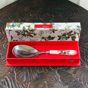 Può includere: Un cucchiaio da portata in metallo argentato con manico in ceramica bianca decorato con un motivo botanico di bacche rosse e foglie verdi. Il cucchiaio è presentato in una scatola rettangolare con interno rosso e coperchio decorato.