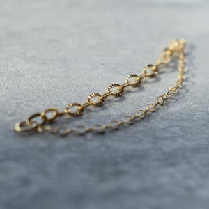 May include: A delicate gold-coloured chain necklace with a series of small, circular links and decorative, scalloped accents. The necklace is shown against a neutral grey background, highlighting its intricate design and craftsmanship. The chain is approximately 18 cm long.