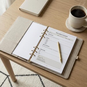 Può includere: Un'agenda beige con una penna d'oro su un tavolo di legno. L'agenda è aperta su una pagina intitolata "DAILY EXECUTION PAGE". Una tazza di caffè è appoggiata su un sottobicchiere accanto all'agenda. C'è anche un taccuino sul tavolo.
