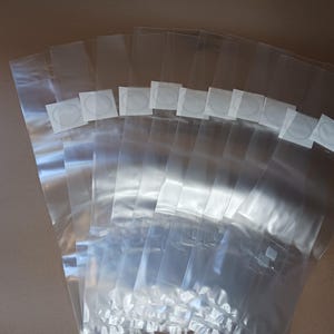 May include: A collection of clear cellophane bags, fanned out to display their contents. Each bag has a small, square white sticker with a circular design. The bags are arranged on a neutral-toned surface.