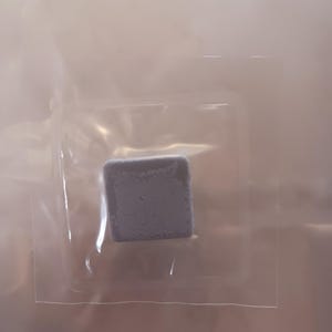 May include: A small, square, gray object is encased in a clear plastic package. The object appears to be a solid, possibly a block or a tablet. The packaging is transparent, allowing the object to be fully visible.
