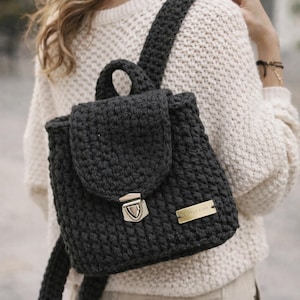 May include: A dark gray crochet backpack with a flap closure and a metal clasp. The backpack has adjustable straps and a small handle. A gold-colored rectangular tag is attached to the front.