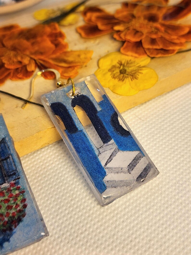 Pu&ograve; includere: Un orecchino rettangolare in resina con un'immagine stampata di una struttura architettonica blu con porte ad arco e gradini in pietra bianca. L'orecchino &egrave; fissato a un gancio in metallo dorato e presenta una finitura lucida.