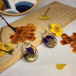 Può includere: Un paio di orecchini in resina a forma di goccia con fiori viola e fogliame verde incorporati. Ogni orecchino è fissato a un gancio in metallo dorato. Gli orecchini sono esposti accanto a petali di fiori secchi arancioni e gialli.