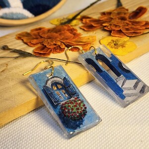 Pu&ograve; includere: Un paio di orecchini pendenti rettangolari con finitura in resina trasparente. Un orecchino raffigura una finestra ad arco blu con fioriera rossa, l'altro una struttura architettonica blu con gradini. Entrambi hanno ganci in metallo dorato.