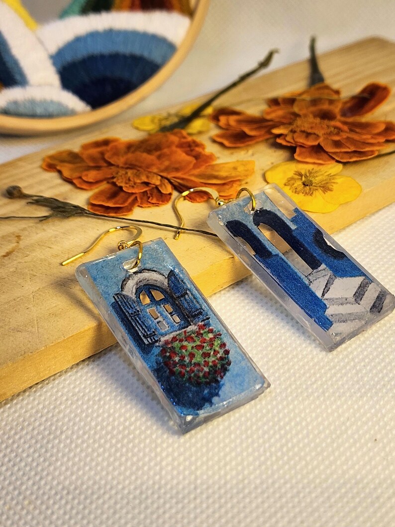 Pu&ograve; includere: Un paio di orecchini pendenti rettangolari con finitura in resina trasparente. Un orecchino raffigura una finestra ad arco blu con fioriera rossa, l'altro una struttura architettonica blu con gradini. Entrambi hanno ganci in metallo dorato.